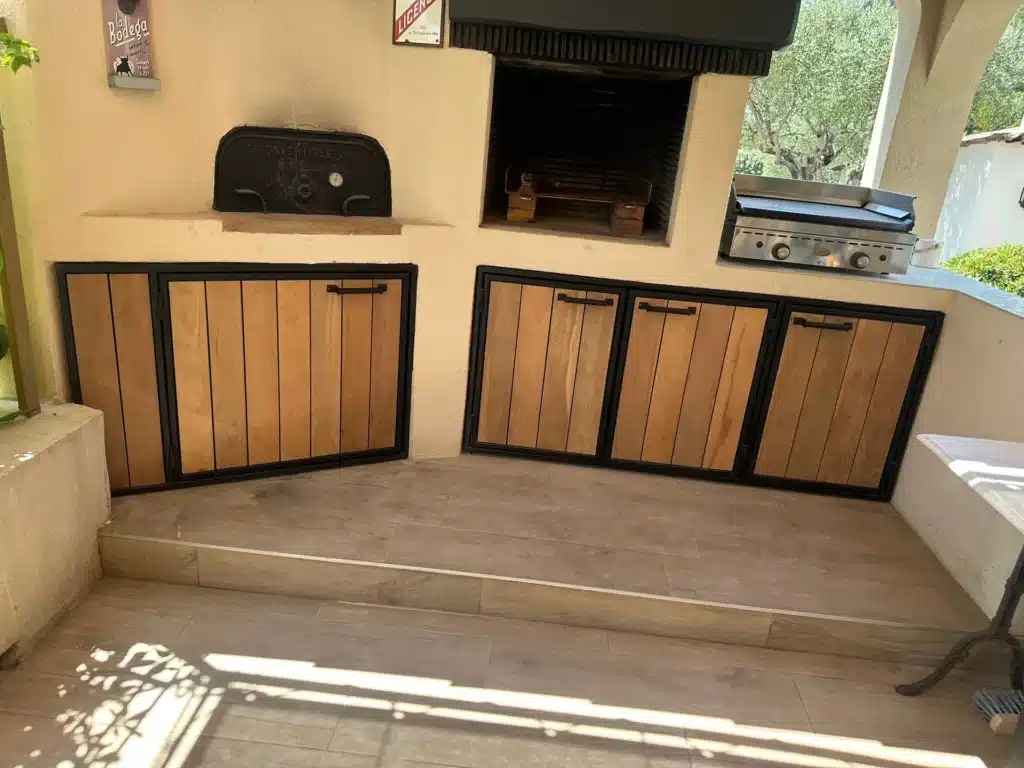 Cuisine d'été rustique avec four à bois et plancha Cuisine d'été rustique avec four à bois Le Convertible, barbecue et placards en bois et métal noir.