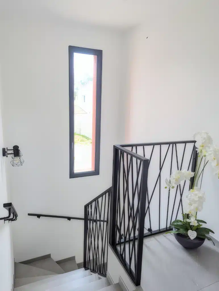 Déco Escalier Moderne : Garde-Corps en Métal Noir Escalier moderne avec garde-corps en métal noir, fenêtre étroite et orchidée blanche dans un intérieur lumineux.