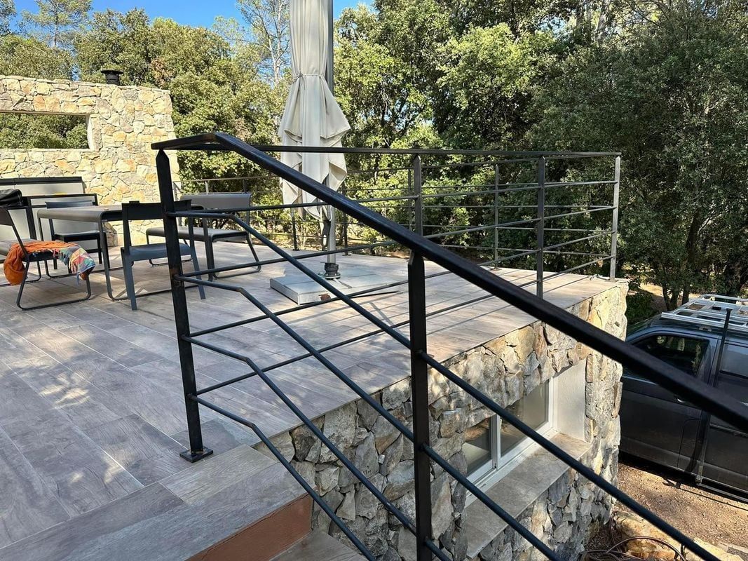 Garde-corps terrasse en fer forgé noir et mur en pierre Terrasse avec garde-corps en fer noir, carrelage effet bois et mur en pierre naturelle sur fond de forêt.